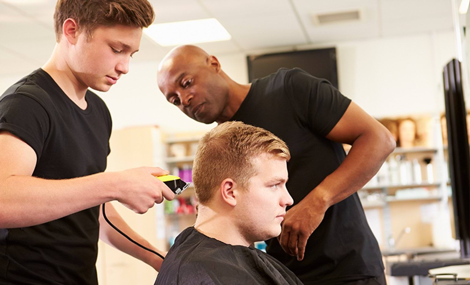 A teenage boy is buzz cutting a man's hair while his teacher assists him. All three of them wear black shirts.