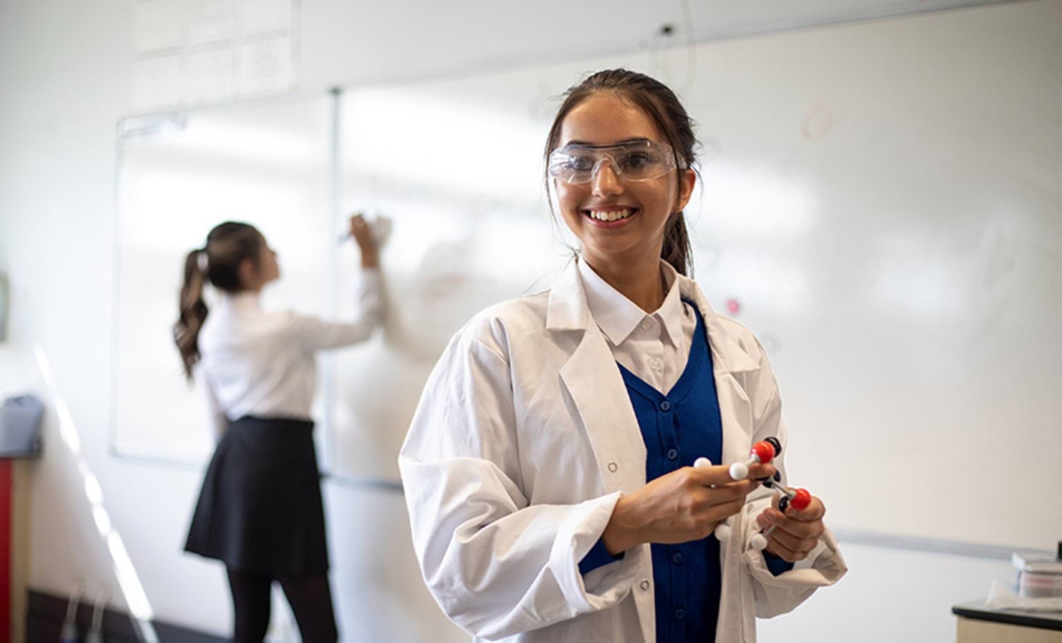 A teenage girl in a lab coat and protective glasses smiles to someone off camera. She is holding a DNA model in her hands.