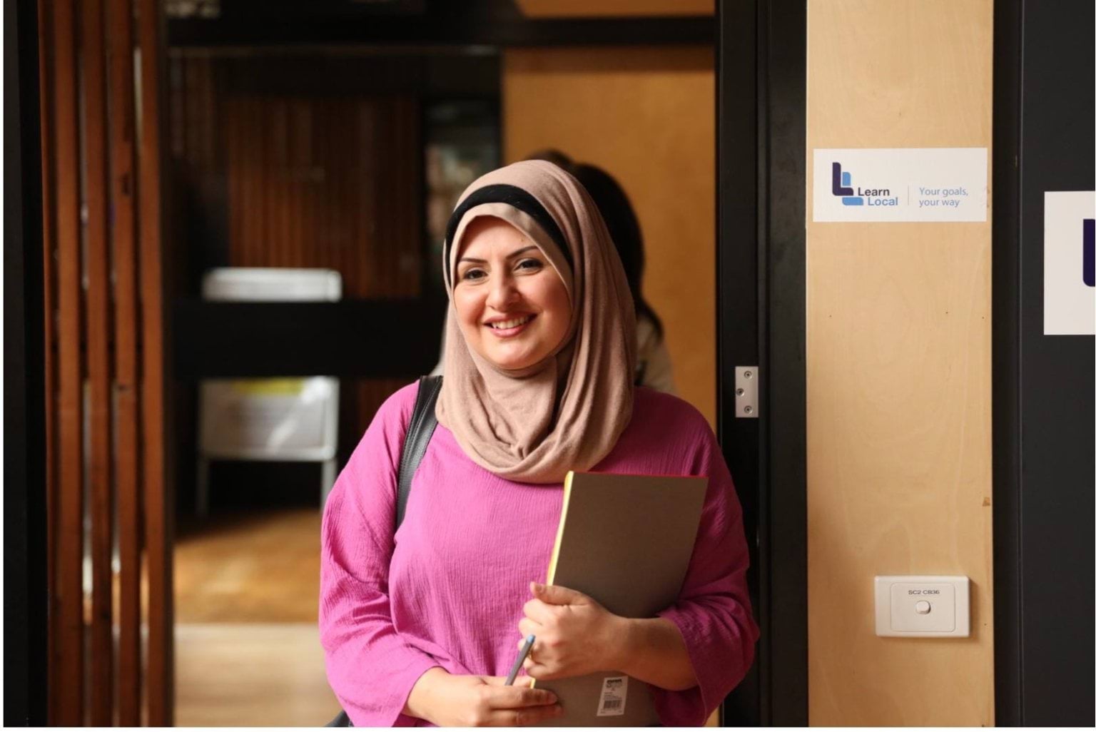 Person in pink top and hijab standing in a doorway with a Learn Local sign on it.