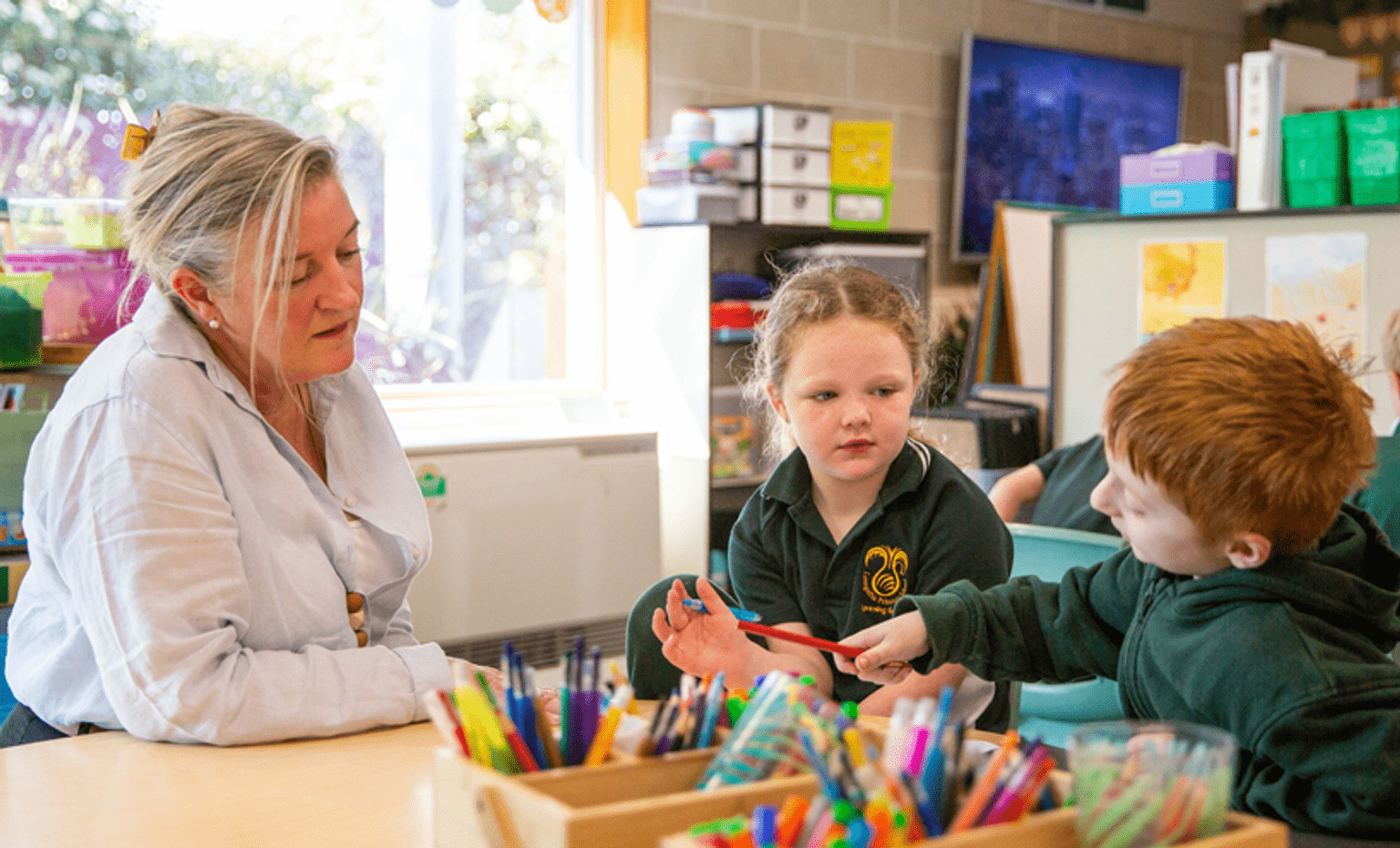 Become a teacher A female teacher talks to two children in green uniforms over a table with lots of colourful pens and pencils.