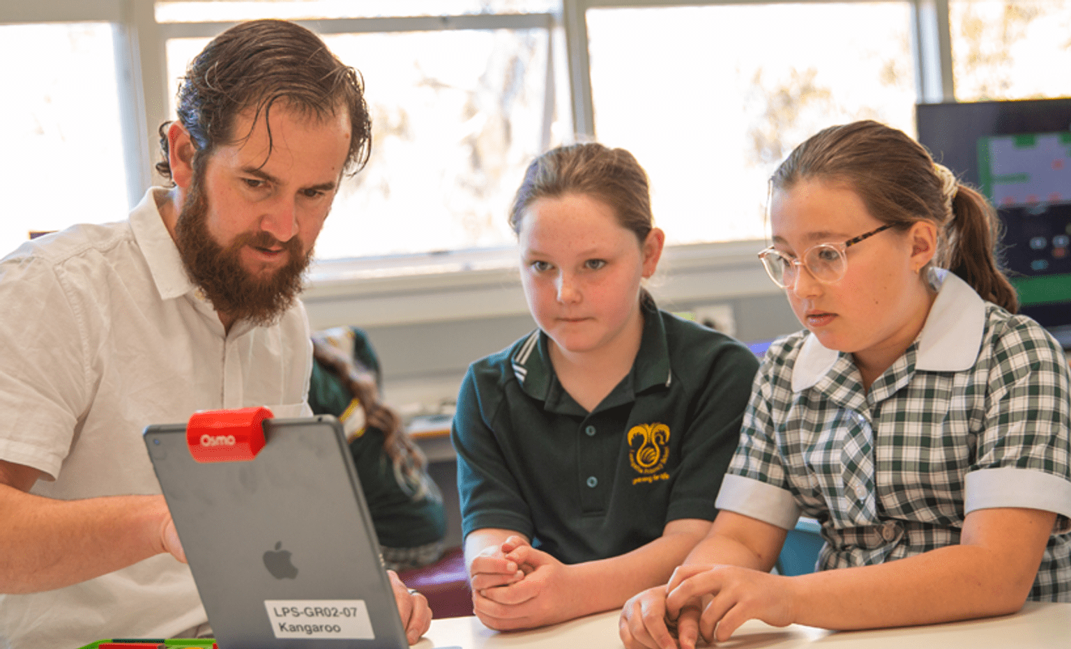 Contact the Department of Education A male school teacher in a white shirt sits with two female students in a green t-shirt and a green and white school dress. They are all looking at an iPad and mid conversation.