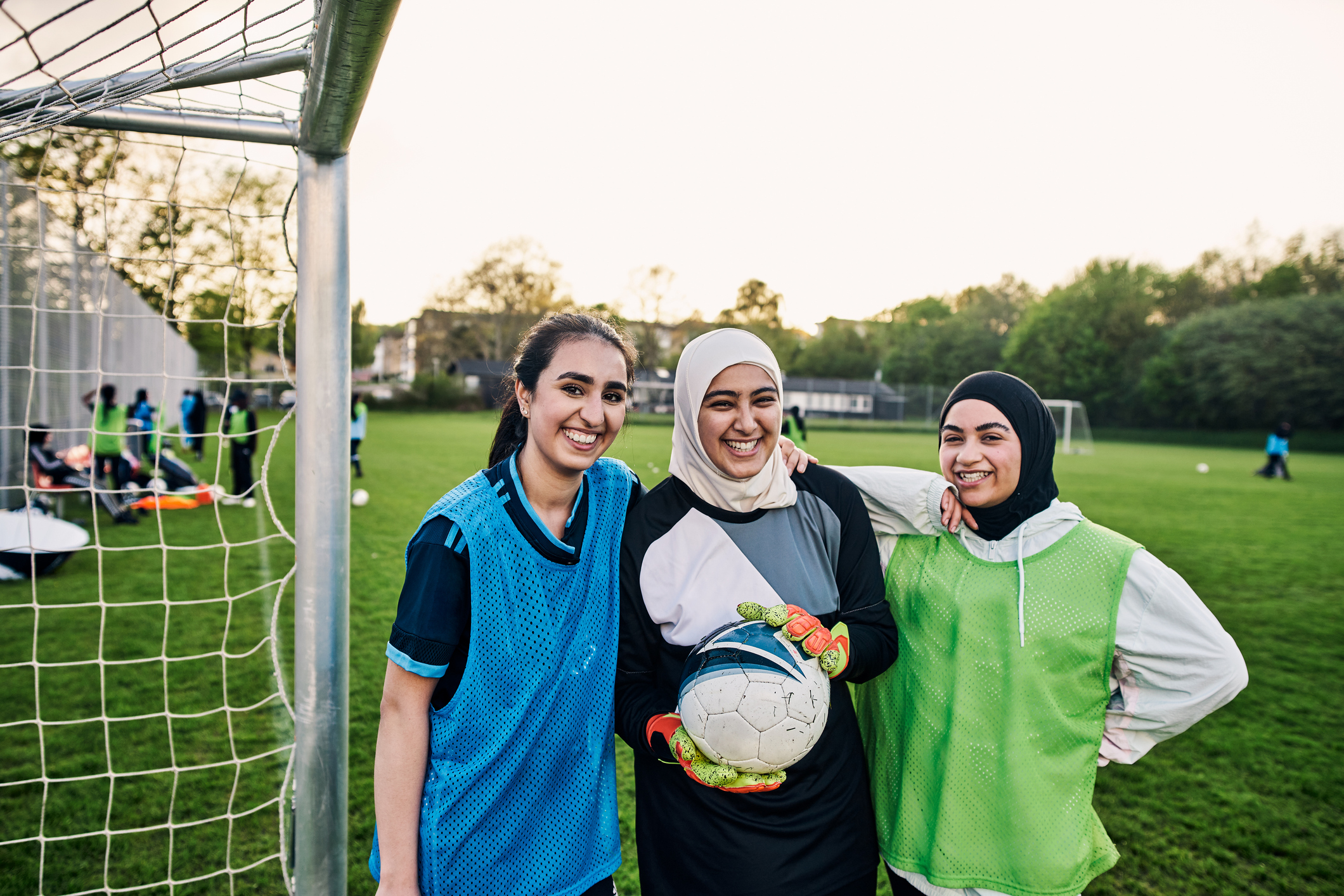 A group of three smiling girls in soccer gear stand next to a goal net. Two of the girls wear Islamic headscarves.