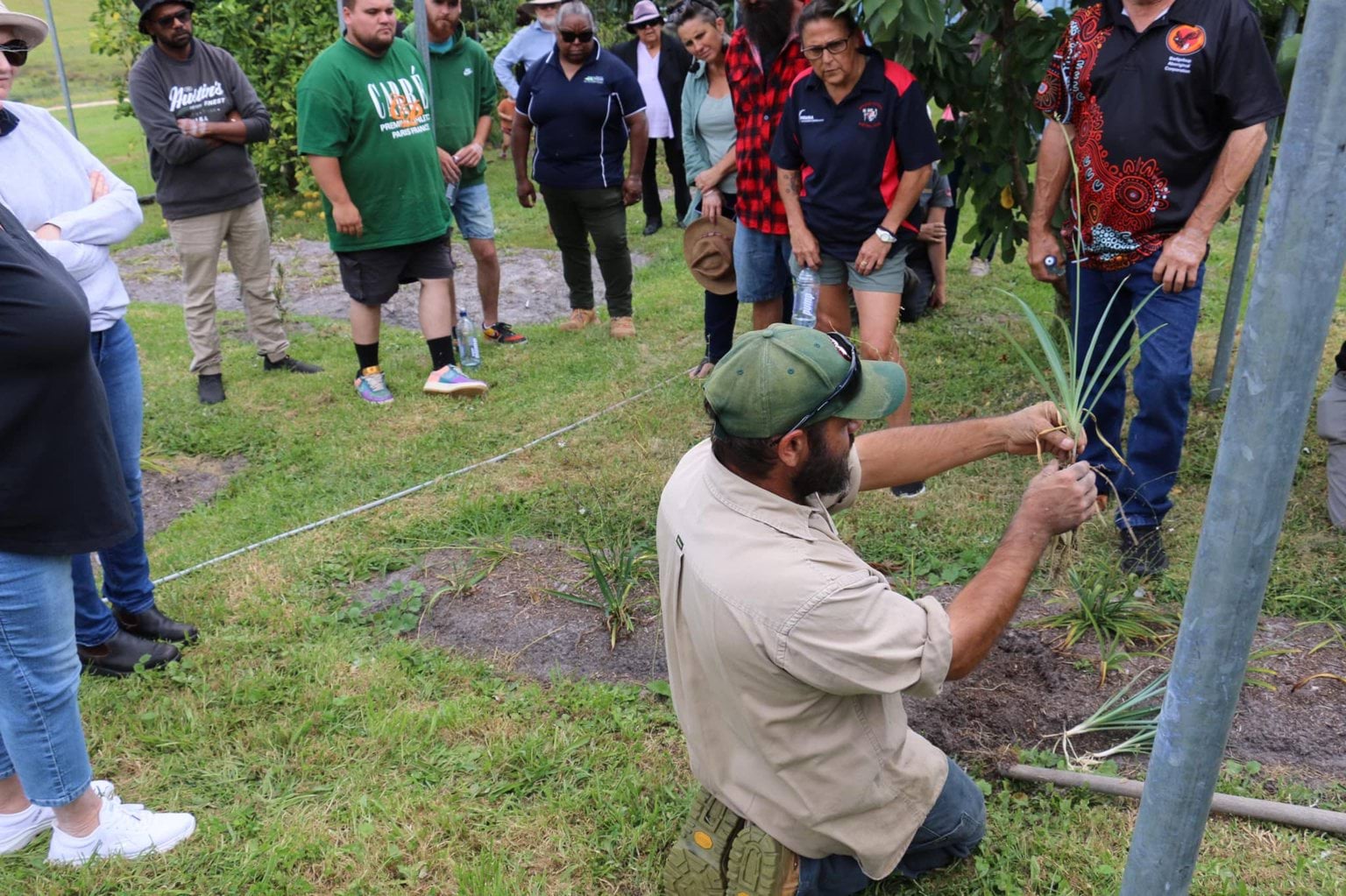 Man in a green hat and brown shirt kneels as he holds a plant in demonstration to a large crowd of people