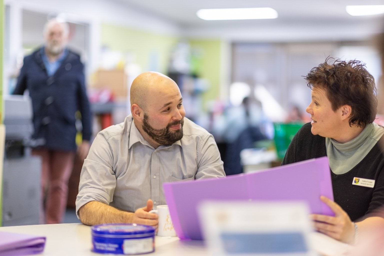 A man and a woman sitting at a table chatting. The woman is holding a purple folder.
