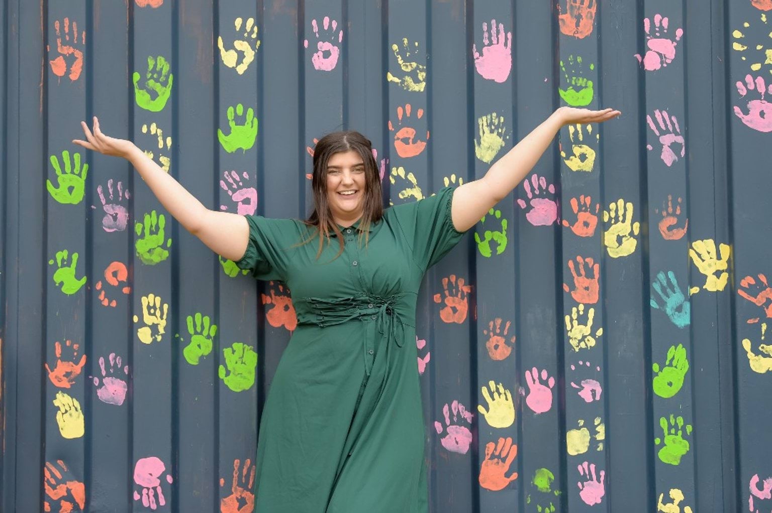 A woman in a green dress stands with arms raised in front of a wall covered with colourful handprints.