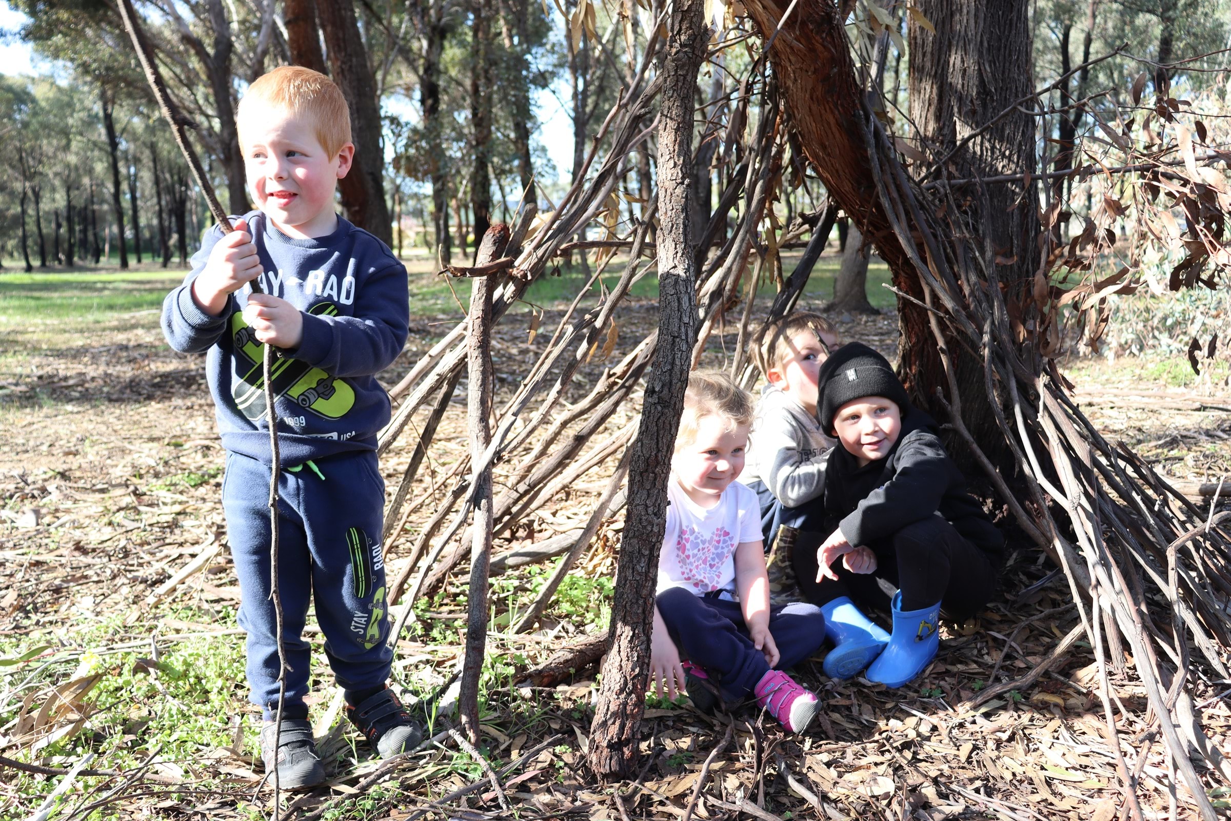 Three young children sitting under a tree in the bush with a young boy holding a stick.   