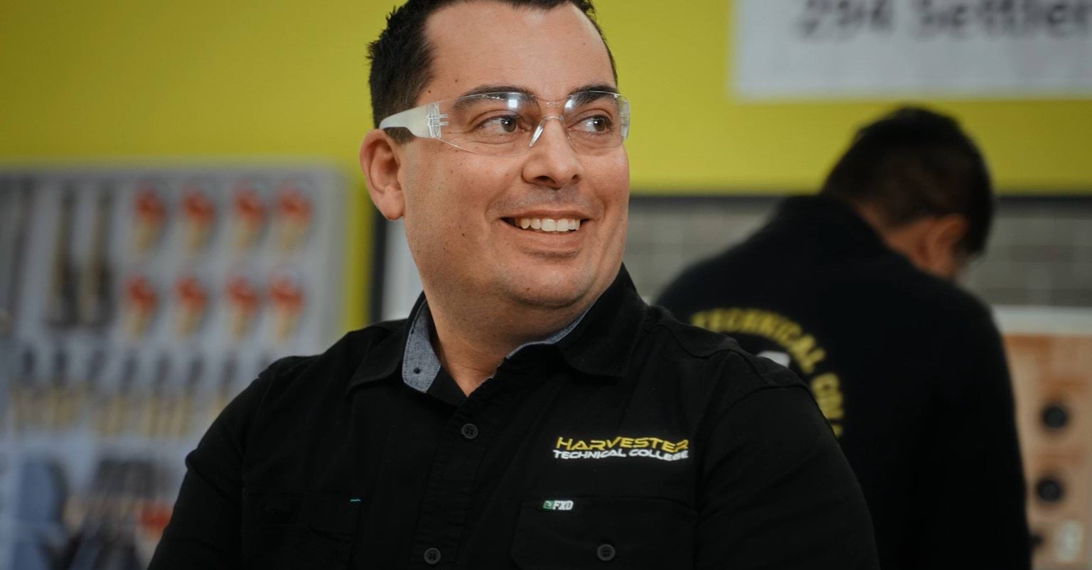A man wearing clear glasses and a black polo shirt, smiling with his head turned sidesways.