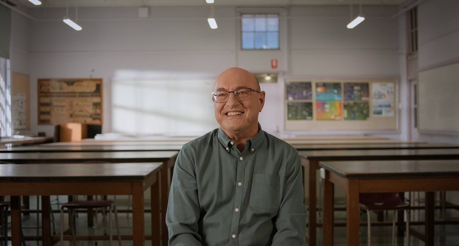 A man wearing glasses and a green button-up shirt sits in a classroom.
