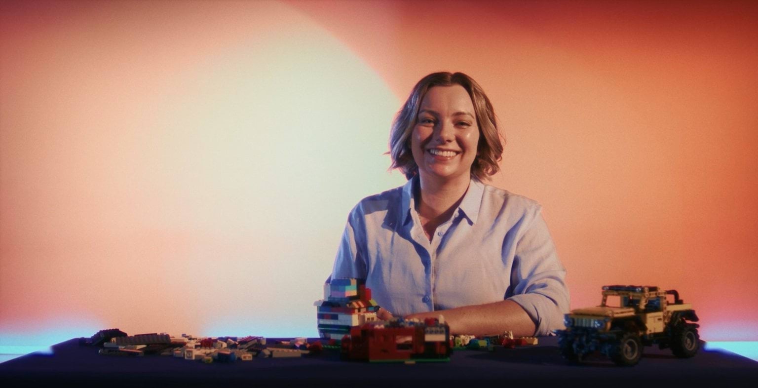 A woman smiling and sitting at a table covered with colourful Lego pieces.