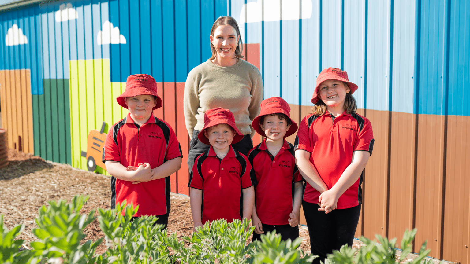 A woman stands behind 4 school children wearing red polo shirts and bucket hats.