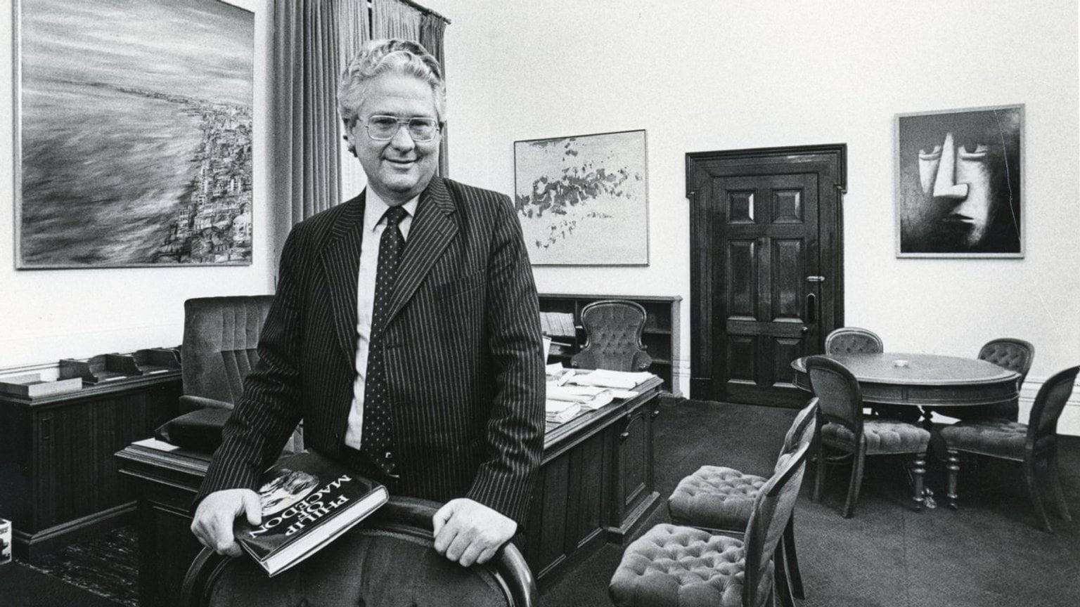 The Honourable Dr Race Mathews The Honourable Dr Race Mathews is wearing a pin-stripe suit and glasses. He leans on the back of a chair in his office holding a book titled Philip of Macedon