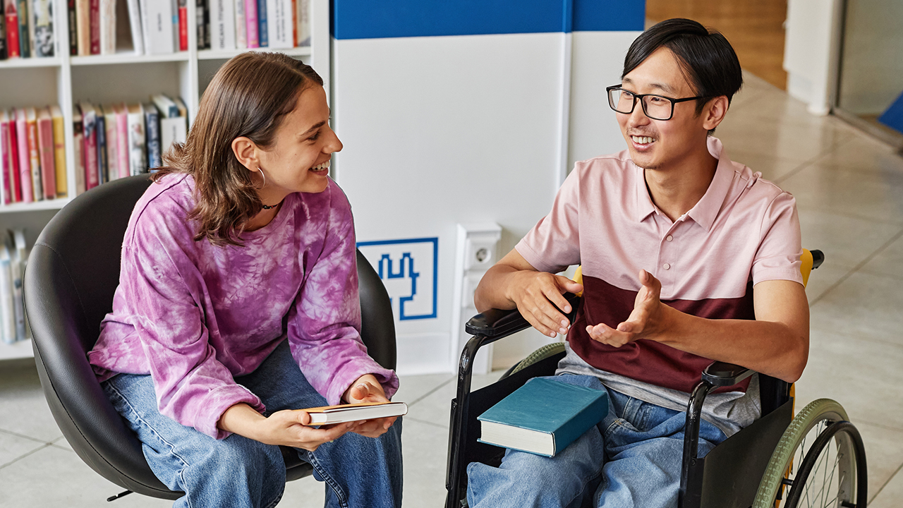 Two people talking in a library setting one person is using a wheelchair