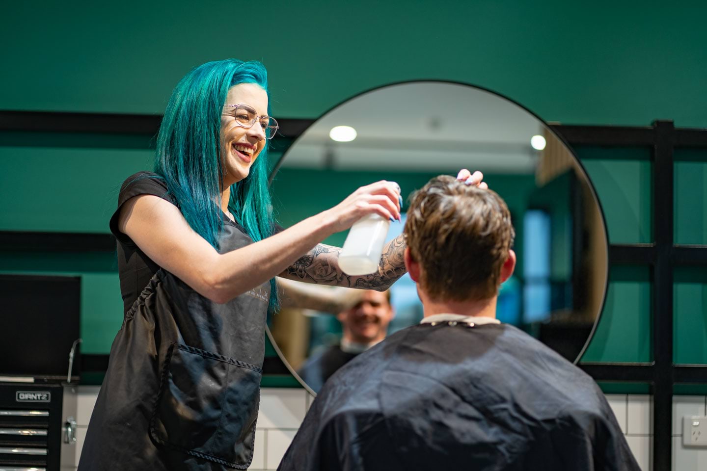 A person in a black apron sprays water on a person sitting in a chair facing a mirror.