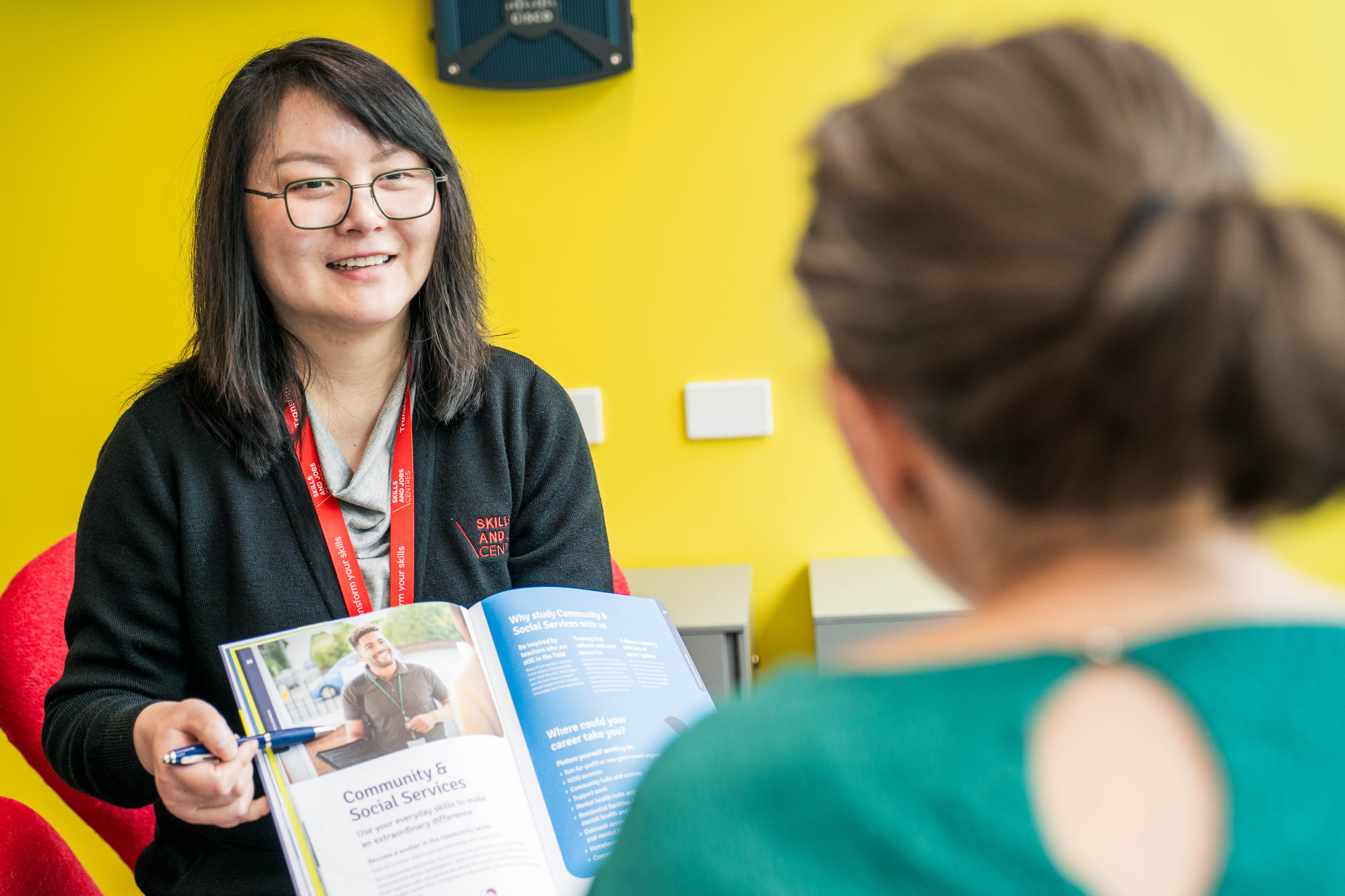 A smiling person wearing glasses and a red lanyard shows a book to a person with their back to the camera.