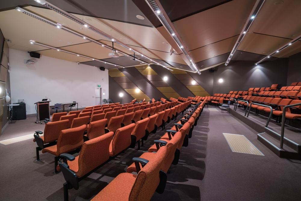 An auditorium with bright lighting, rows of orange chairs and grey carpet.