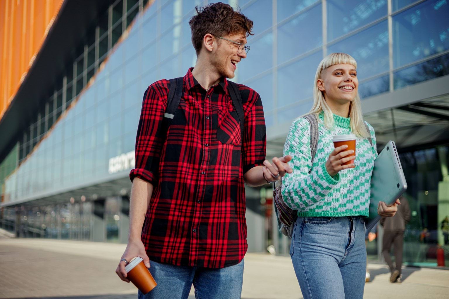 Two people carrying coffee cups smiling and walking past a glass building