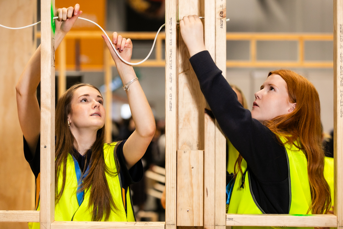 Two female students helping each other run cable through a wooden house frame