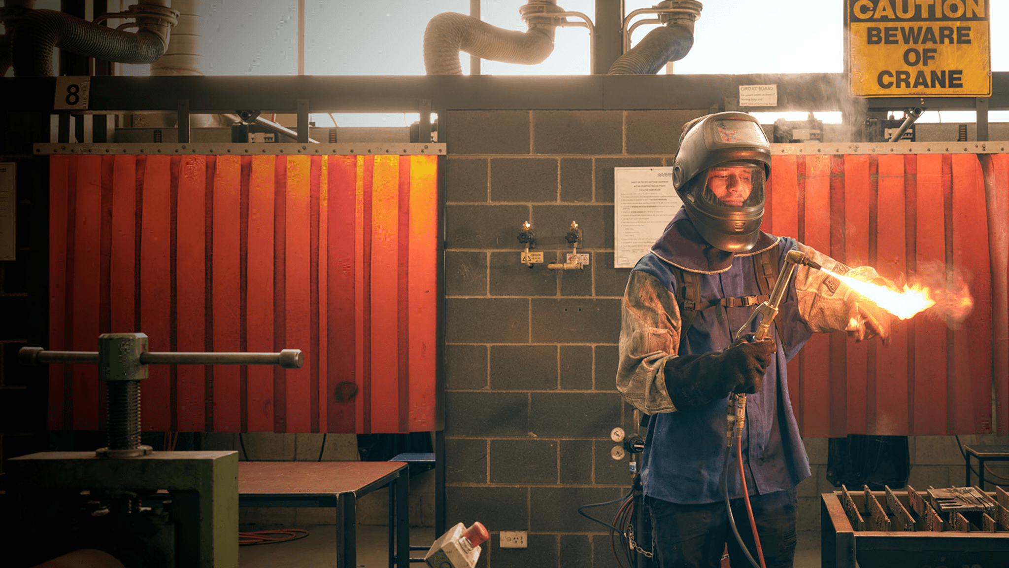 A person in welding gear and helmet lights a torch in an industrial setting.