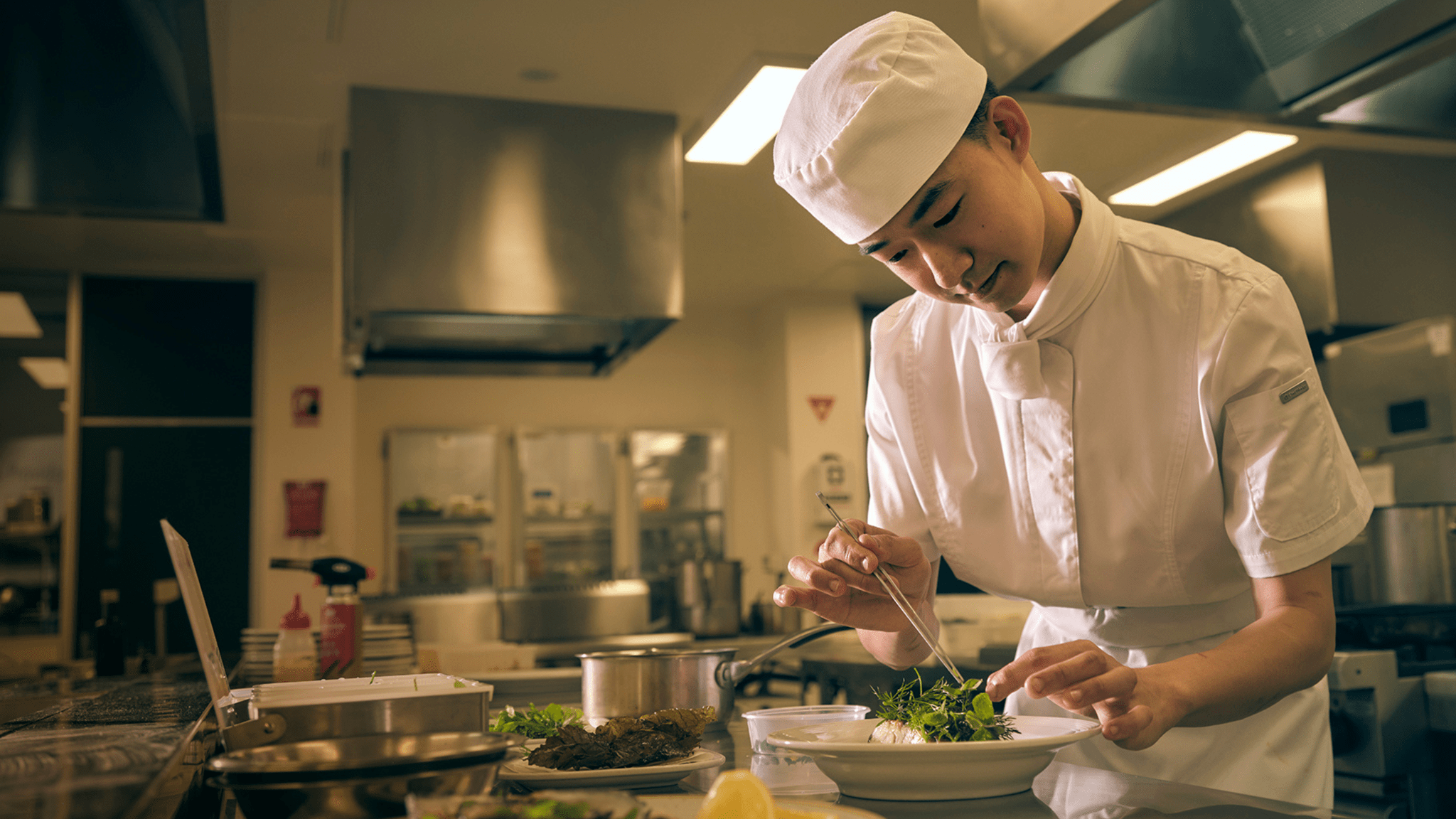 A person in a chef's uniform and apron uses tweezers to place food on a plate in a commercial kitchen.