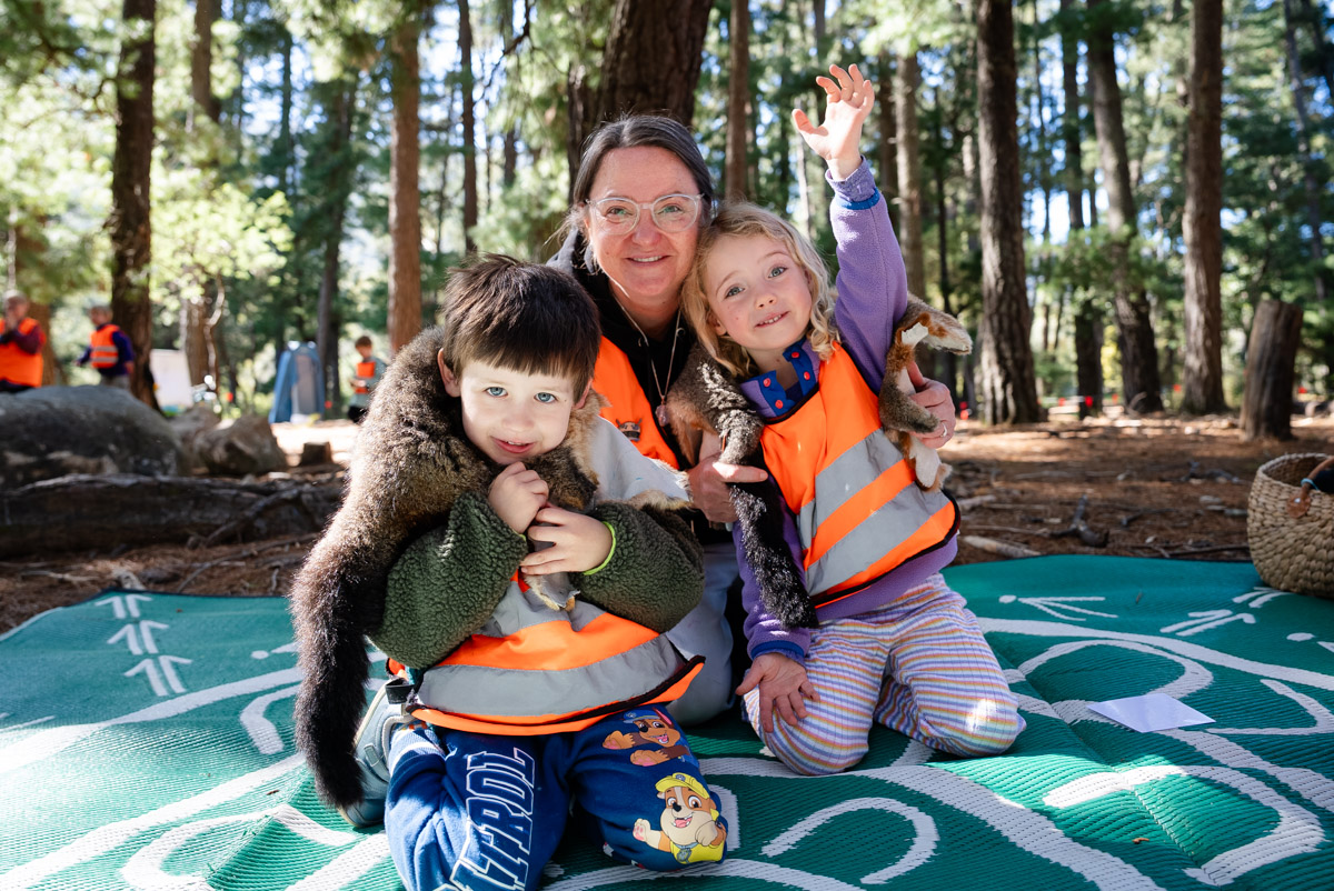 Teacher and 2 small children kneeling on a blanket in the bush.