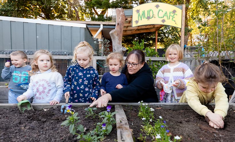 teacher and children planting flowers in a garden bed.