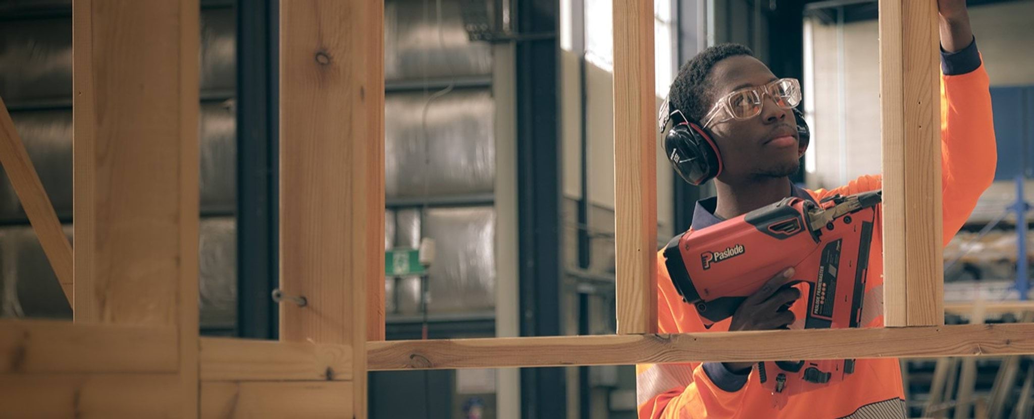 A person in hi-vis, headphones and safety glasses uses a nail gun on a wooden frame.