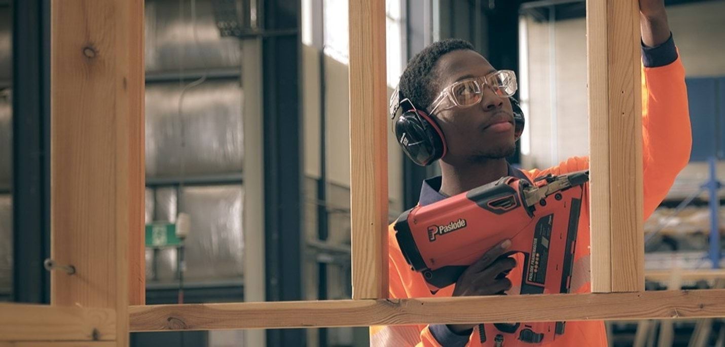 A person in hi-vis, headphones and safety glasses using a nail gun on a wooden frame.