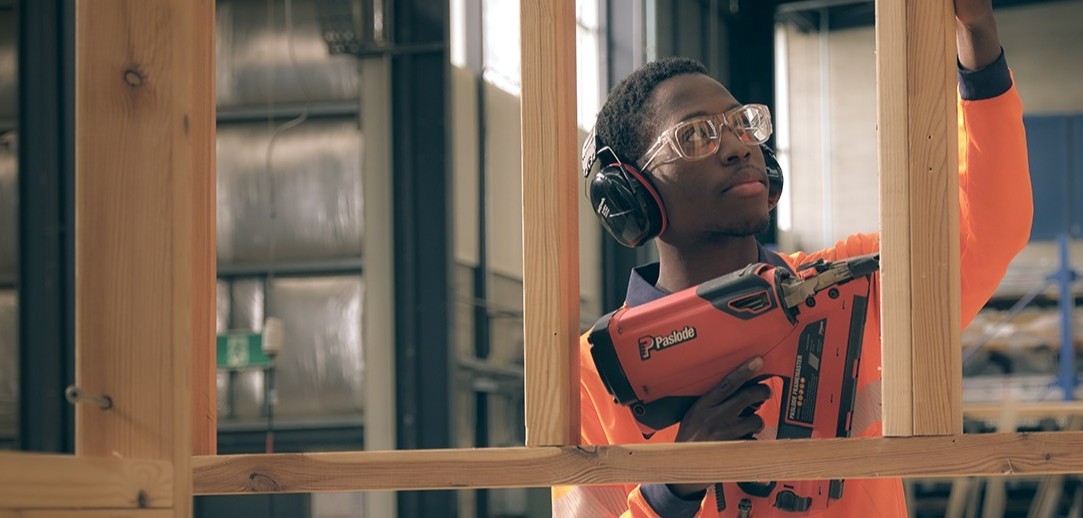 A person in hi-vis, headphones and safety glasses using a nail gun on a wooden frame.